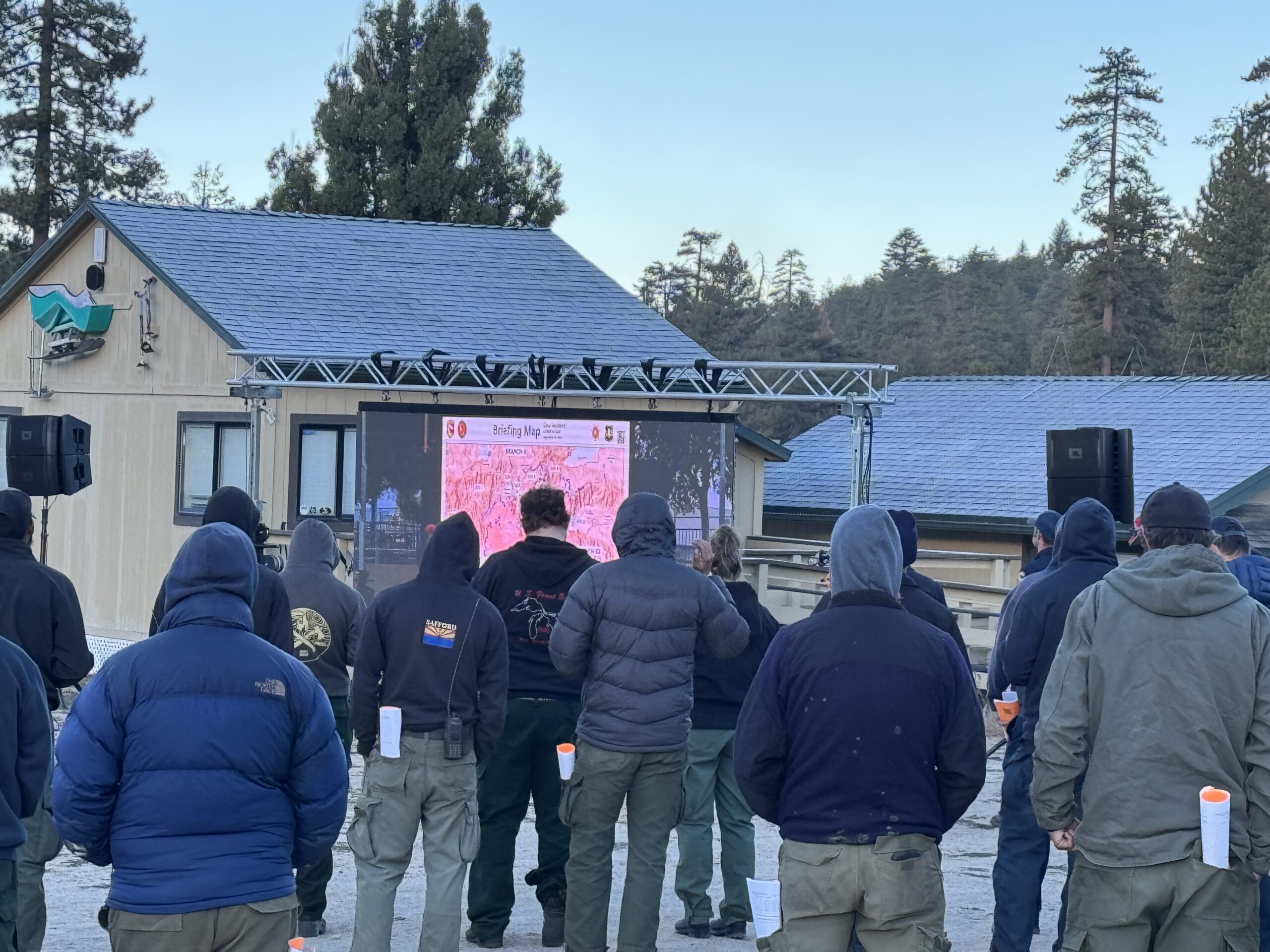 Fire crews at a morning operational briefing viewing a briefing map on a base-camp LED display with line-array speakers flanking the screen.